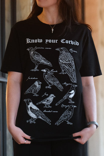 A person wearing a black short-sleeve graphic T-shirt with white bird illustrations and the text 'Know your Corvid' across the chest.