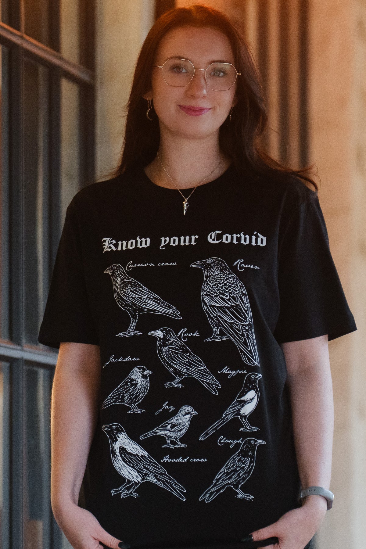 A woman wearing a black graphic t-shirt with white line drawings of various corvid birds and the text 'Know your Corvid' printed on the front, gothic bookish style