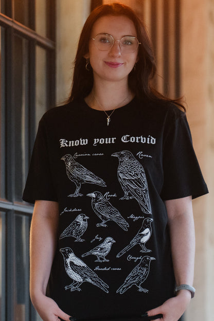 A woman wearing a black graphic t-shirt with white line drawings of various corvid birds and the text 'Know your Corvid' printed on the front, gothic bookish style