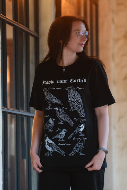 A woman wearing a black t-shirt with a white graphic print of various corvid birds and the text 'Know your Corvid' on the front.